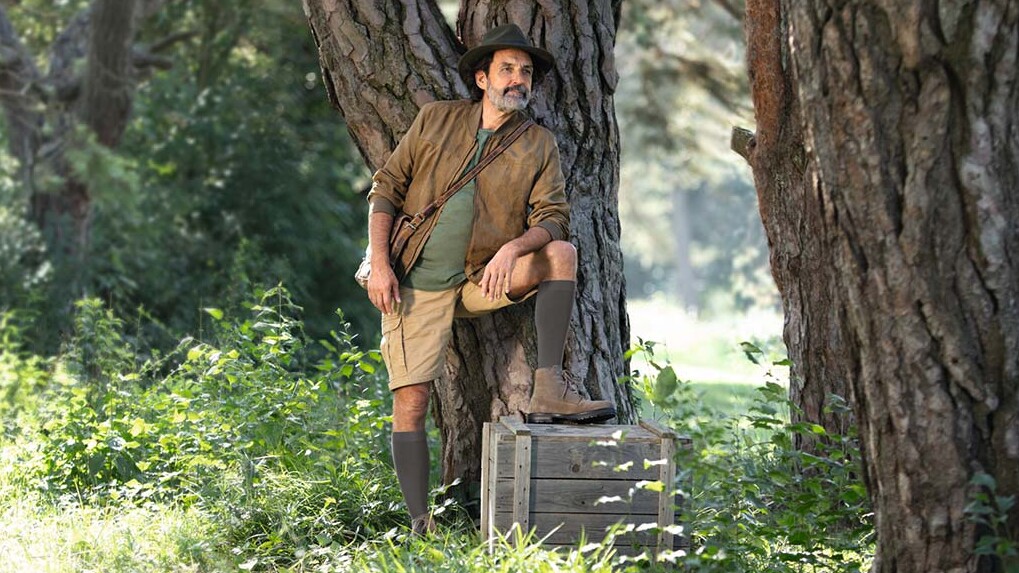 Ein Mann mit Entdecker-Outfit steht in der Natur und schaut in die Ferne. Er trägt kniehohe Kompressionsstrümpfe.