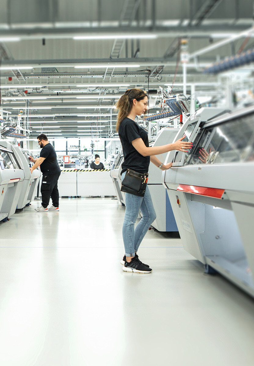 Person bedient eine Flachstrickmaschine in der Produktionshalle von Juzo, während weitere Personen im Hintergrund an ähnlichen Maschinen arbeiten.