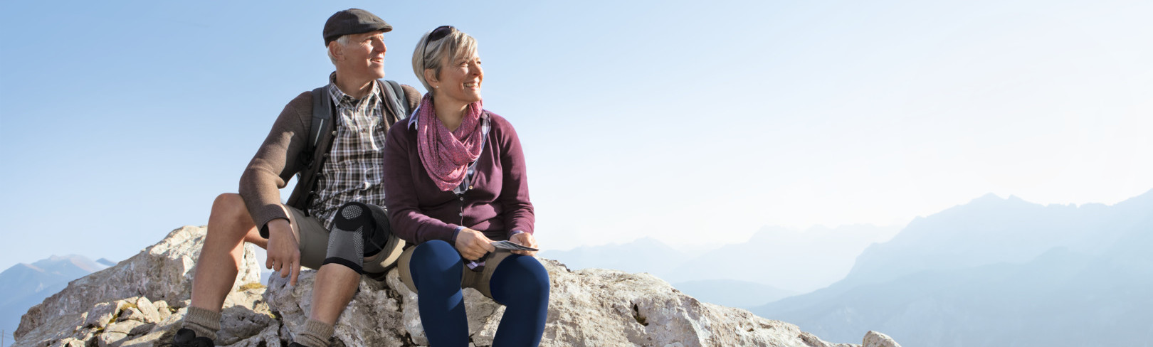 Lifestylebild Wandern Mann mit Kniebande Genu Xtra und Frau mit Kompressionsstrumpf Juzo Dynamic sitzen auf einem Felsen und blicke´n in die Ferne