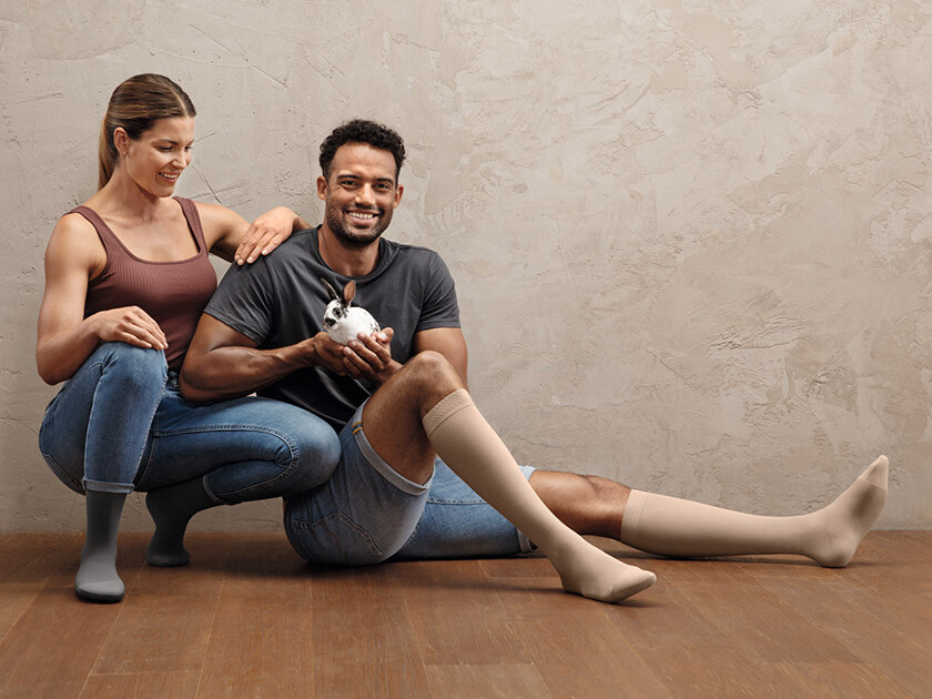 Juzo Intenso A man and a woman are sitting on the floor. Both are wearing compression stockings on their lower legs. The man is holding a small bunny in his hands
