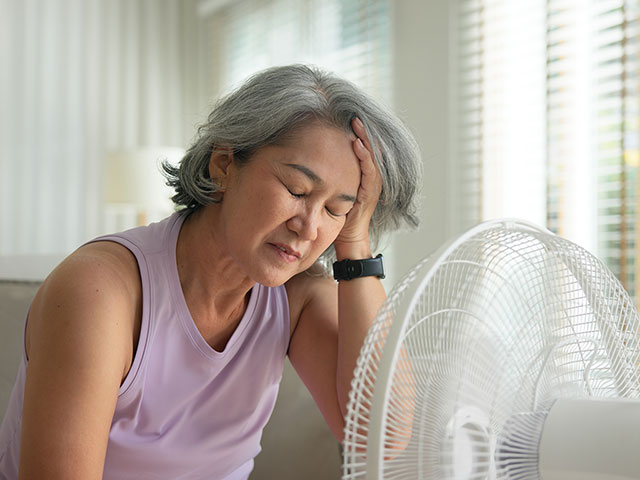 Ältere Frau in den Wechseljahren hat Kopfschmerzen und sitzt vor einem Ventilator