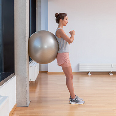 Una mujer se apoya contra la pared con la espalda recta, apoyada en una pelota de gimnasia