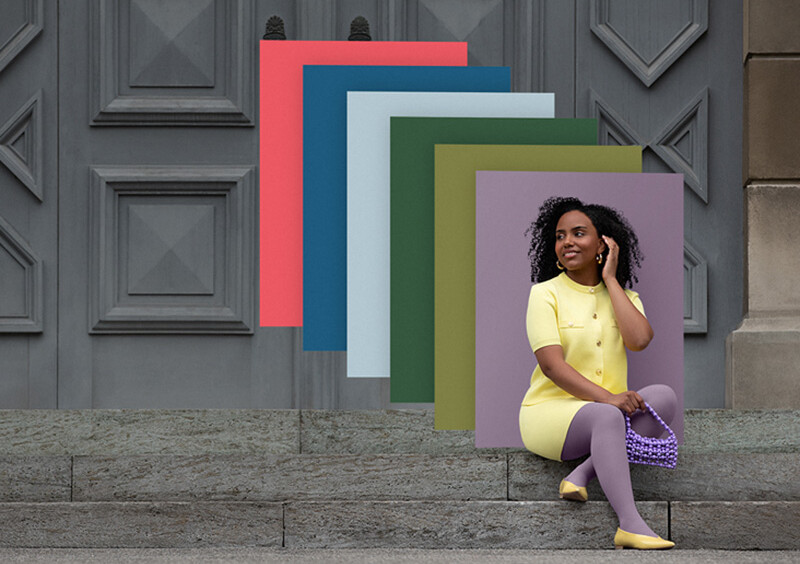 En kvinde sidder på stentrapper foran en port. Hun bærer et lysegult todelt sæt til en lilla kompressionsbandage. Bag hende ses farvefelter i trendfarverne.