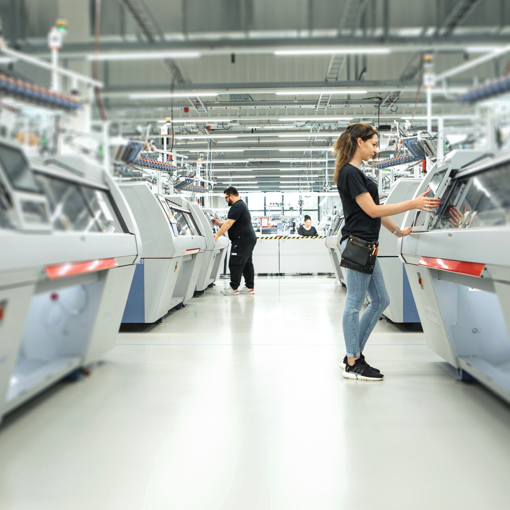Person bedient eine Flachstrickmaschine in der Produktionshalle von Juzo, während weitere Personen im Hintergrund an ähnlichen Maschinen arbeiten.