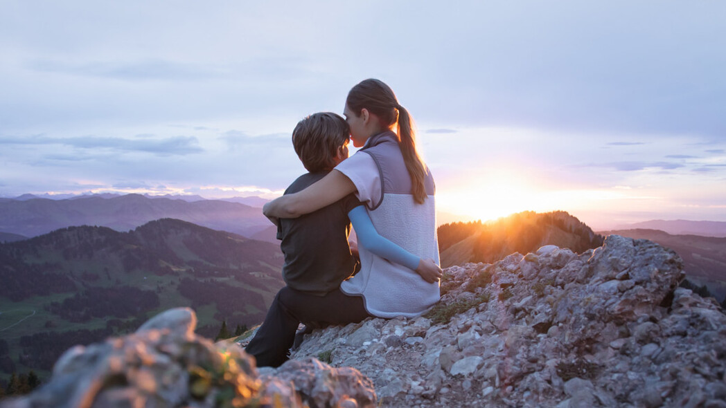 A mother is sitting on a mountain peak with her child. The child is wearing a compression sleeve. A mother is sitting on a mountain peak with her child. The child is wearing a compression sleeve.