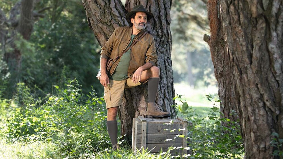 A man dressed in explorer gear is standing in the great outdoors, gazing into the distance. He is wearing knee-high compression socks.