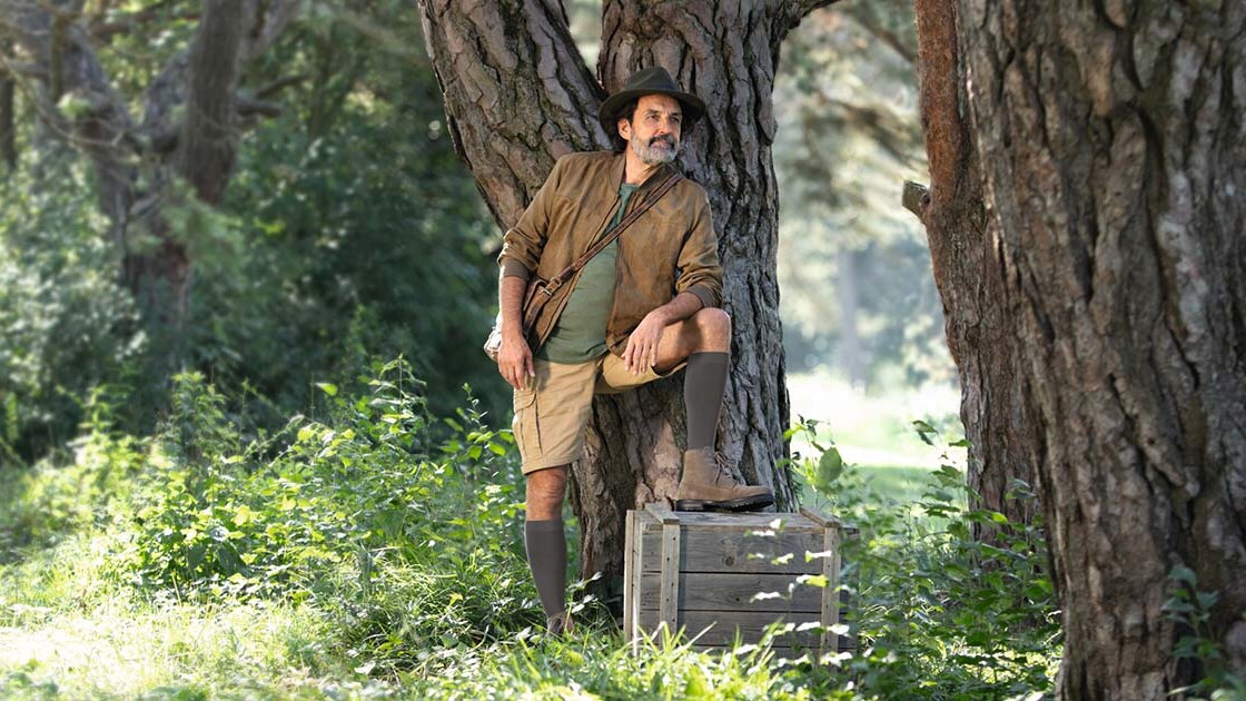 Um homem com um fato de explorador está na natureza e olha para longe. Tem calças de compressão até ao joelho.