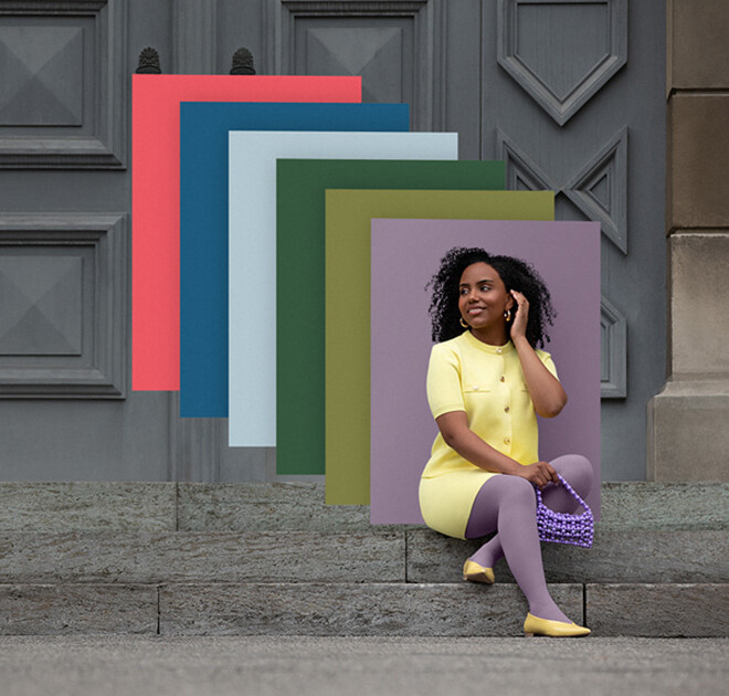 Juzo Trend Colours Batique Une femme est assise sur des marches en pierre devant un portail. Elle porte un ensemble deux pièces jaune clair et un bandage compressif violet. Derrière elle, on aperçoit des aplats de couleurs tendance.