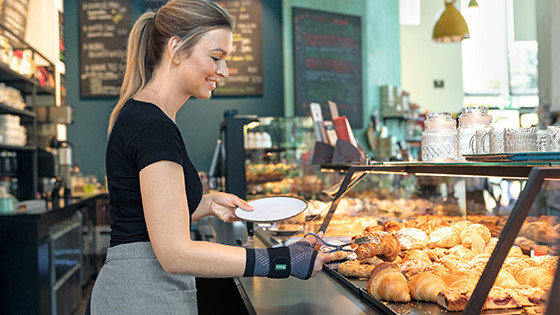 Mainteaser smärtor i handleden (innehållssidor) Bageriexpedit bär handledsbandaget JuzoFlex Manu Xtra och tar en croissant med en serveringstång