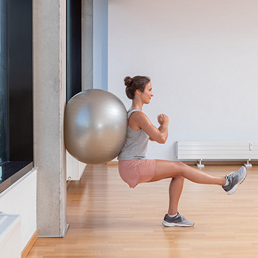 Exercise Wall-sit Step 2 A woman leaning against a wall with an exercise ball, doing a squat with one leg extended