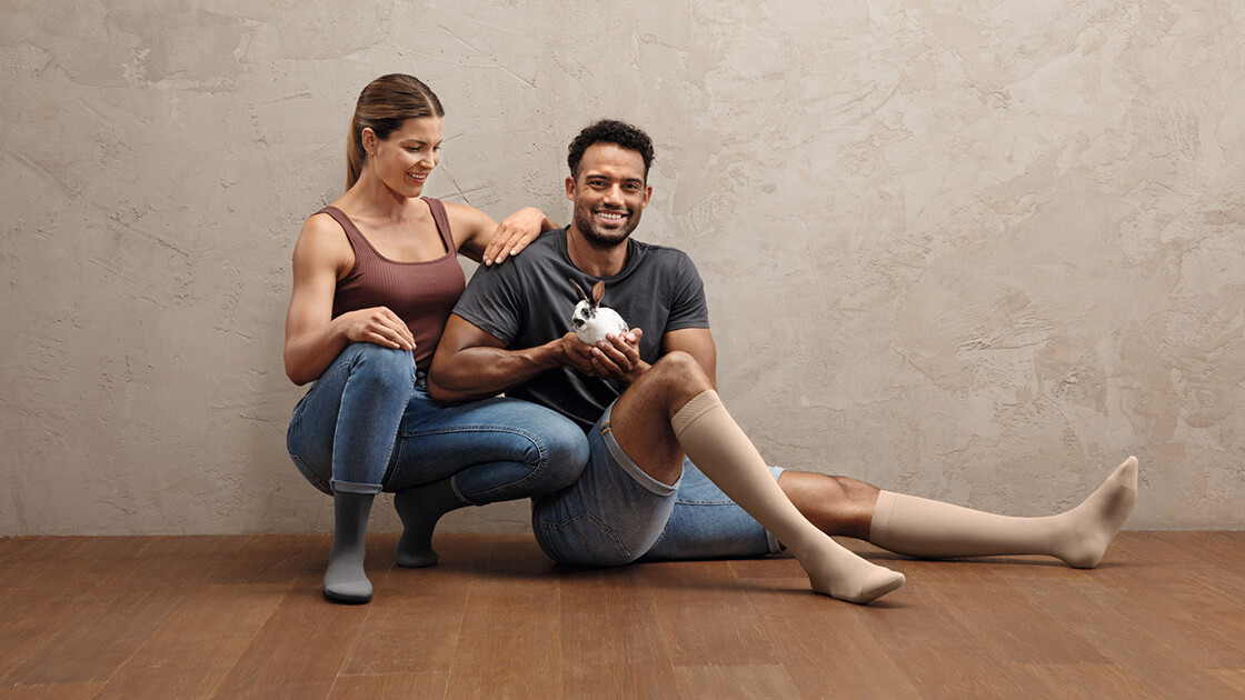 Um casal desportista está agachado no chão. Ambos usam meias de compressão Juzo Intenso. O homem tem um coelhinho na mão. Um casal desportista está agachado no chão. Ambos usam meias de compressão Juzo Intenso. O homem tem um coelhinho na mão.