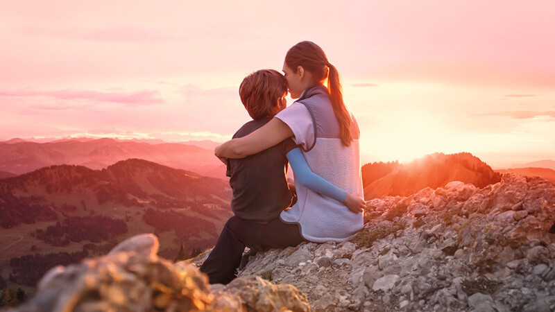 A mother is sitting on a mountain peak with her child. The child is wearing a compression sleeve. A mother is sitting on a mountain peak with her child. The child is wearing a compression sleeve.