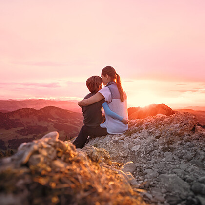 Eine Mutter sitzt mit ihrem Kind im Arm auf einem Berggipfel und betrachtet den Sonnenuntergang