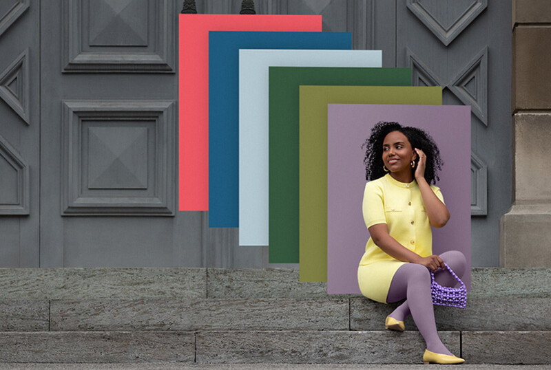 Eine Frau sitzt auf Steinstufen vor einem Tor. Sie trägt einen hellgelben Zweiteiler zu einer lilanen Kompressionsversorgung. Hinter ihr sieht man Farbfelder in den Trend Colours