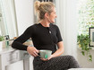 Kvinna med svart thoraxbandage sitter vid ett sminkbord och håller en kopp te i handen