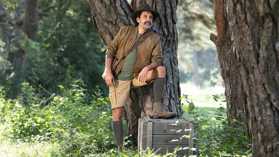 Un homme vêtu d'une tenue d'explorateur se tient en pleine nature et regarde au loin. Il porte des bas de contention qui lui arrivent aux genoux. Un homme vêtu d'une tenue d'explorateur se tient en pleine nature et regarde au loin. Il porte des bas de contention qui lui arrivent aux genoux.