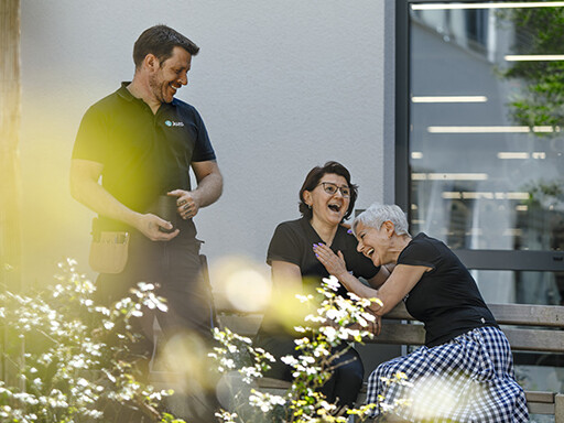 Zwei Frauen mit einem schwarzen Juzo T-Shirt sitzen auf einer Bank und lachen herzhaft, sie unterhalten sich mit einem Mann, der ebenfalls ein schwarzes Juzo T-Shirt trägt und eine schwarze Tasse in der Hand hält