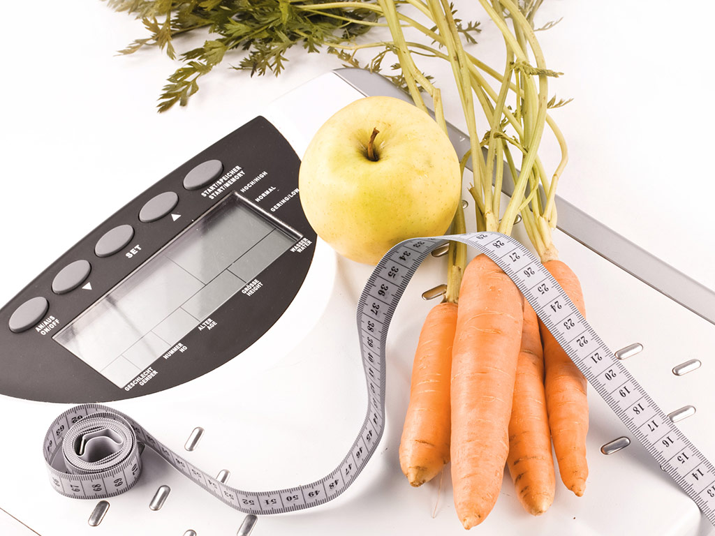 Ein Apfel, ein kleiner Bund Karotten und ein Maßband liegen auf einer Waage