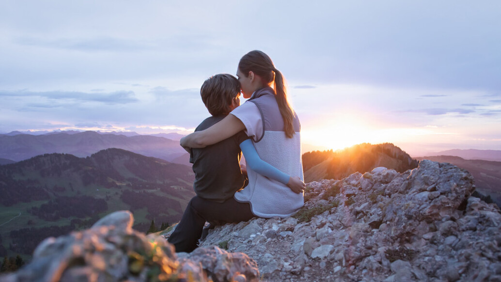 Eine Mutter sitzt mit ihrem Kind auf einem Berggipfel. Das Kind trägt einen Kompressionsärmel. Eine Mutter sitzt mit ihrem Kind auf einem Berggipfel. Das Kind trägt einen Kompressionsärmel.