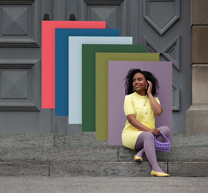 Juzo Trend Colours Batik Een vrouw zit op stenen treden voor een poort. Ze draagt een lichtgele tweedelige outfit met een paarse compressiekous. Achter haar zie je kleurvlakken in de trendkleuren.