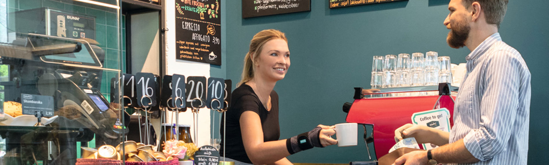 Lifestyle-billede café - JuzoFlex Manu Xtra Ekspedient i bageri bærer håndledsbandagen JuzoFlex Manu Xtra og rækker kunden en kop over disken