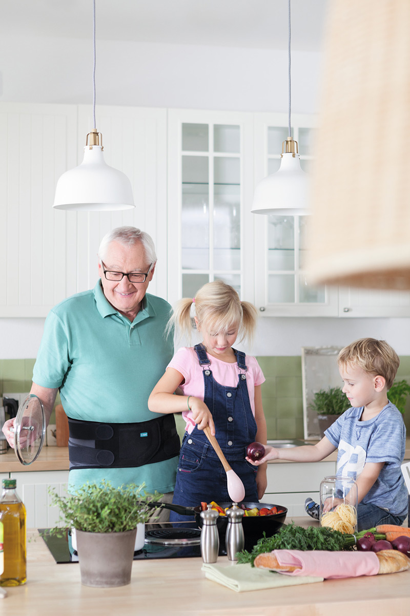 Bedstefar laver mad sammen med sine børnebørn. Han bærer en rygstøtte Bedstefar laver mad sammen med sine børnebørn. Han bærer en rygstøtte
