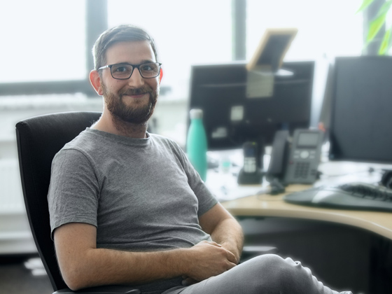 Mitarbeiter in der IT lächelt in die Kamera, während er auf einem Bürostuhl sitzt
